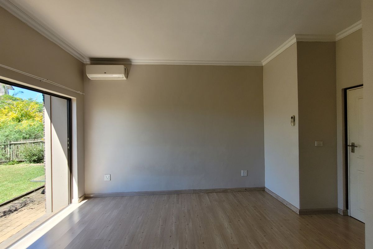 A bright, empty living room featuring light wood-look flooring, crown molding, a wall-mounted air conditioning unit, and sliding glass doors leading to the garden.
