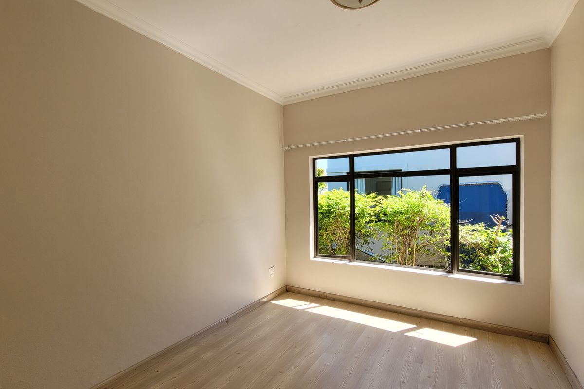 A bright, empty bedroom featuring light-toned laminate flooring, neutral walls, crown molding, and a large window with dark frames overlooking greenery.