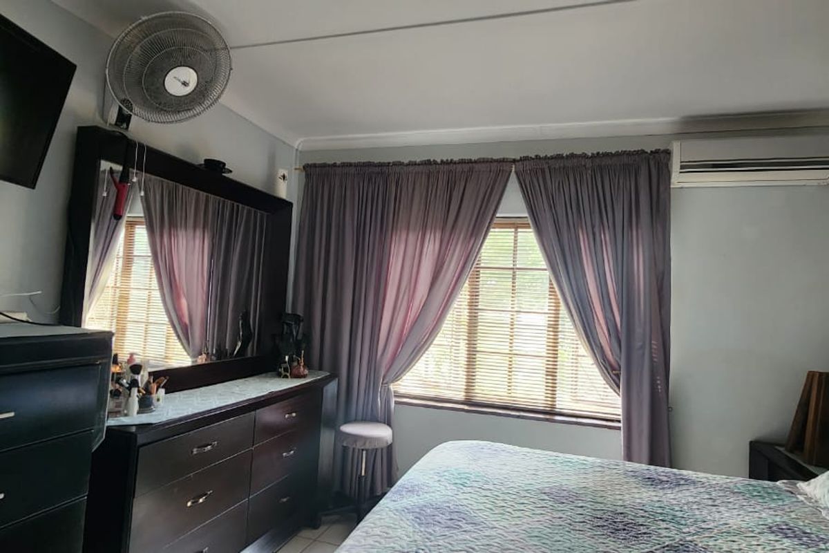 A functional bedroom featuring a wall-mounted air conditioning unit, ceiling fan, and window with fitted blinds and curtains.