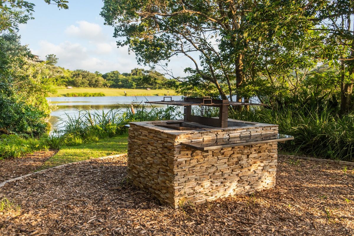 A stone-clad outdoor braai area situated in a scenic garden setting, overlooking a tranquil lake and natural landscape.