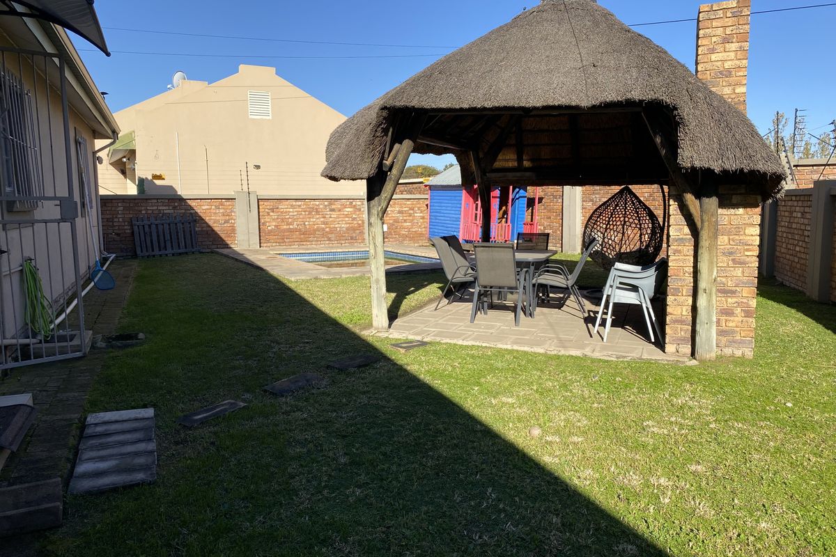 Rear garden featuring a thatched lapa with a brick chimney, paved patio area, and a swimming pool enclosed by brick boundary walls.