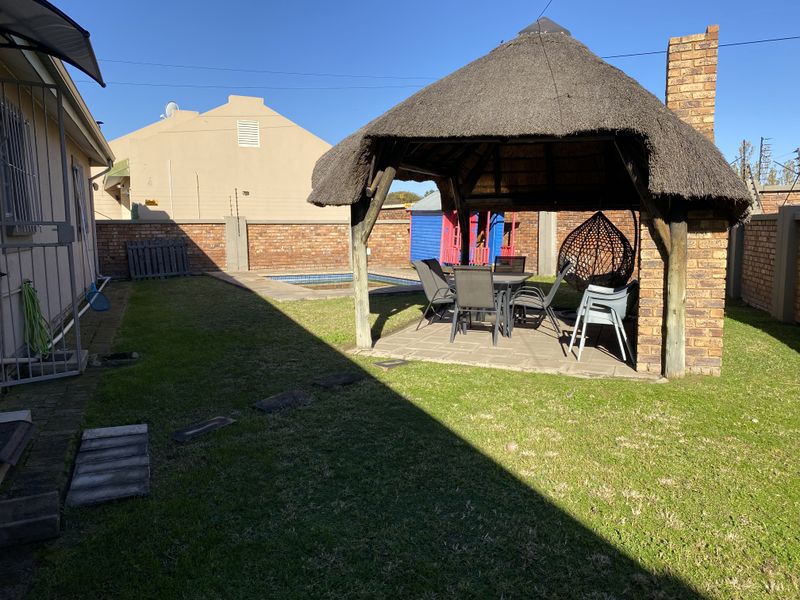 Rear garden featuring a thatched lapa with a brick chimney, paved patio area, and a swimming pool enclosed by brick boundary walls.