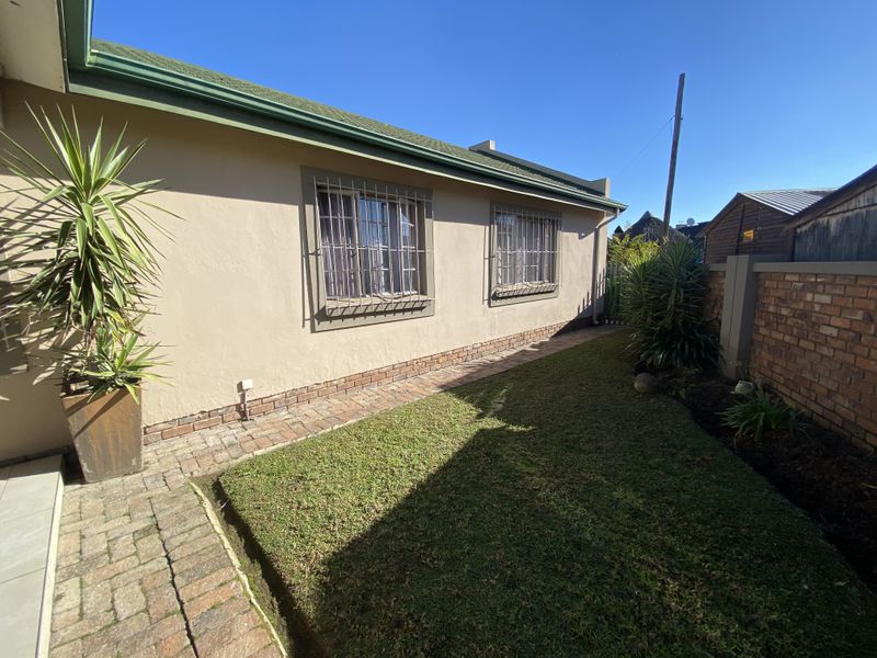 A well-maintained rear exterior view featuring a paved walkway, a small lawn, and secure windows with fitted security bars.