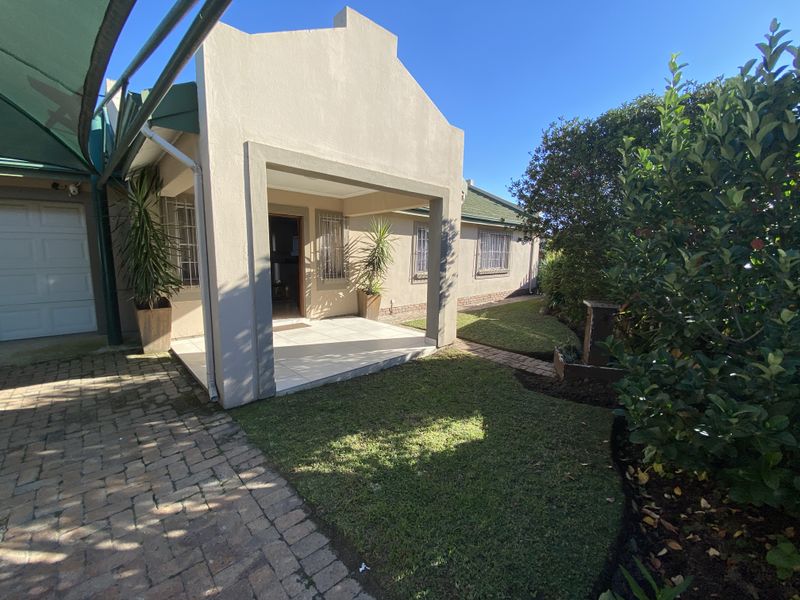 A well-maintained rear exterior view featuring a covered tiled patio, paved driveway, and a neat landscaped garden with security bars on windows.