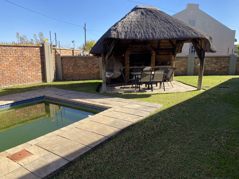 Outdoor entertainment area featuring a thatched lapa with a built-in braai, overlooking a swimming pool with paved surround and enclosed by brick walls.