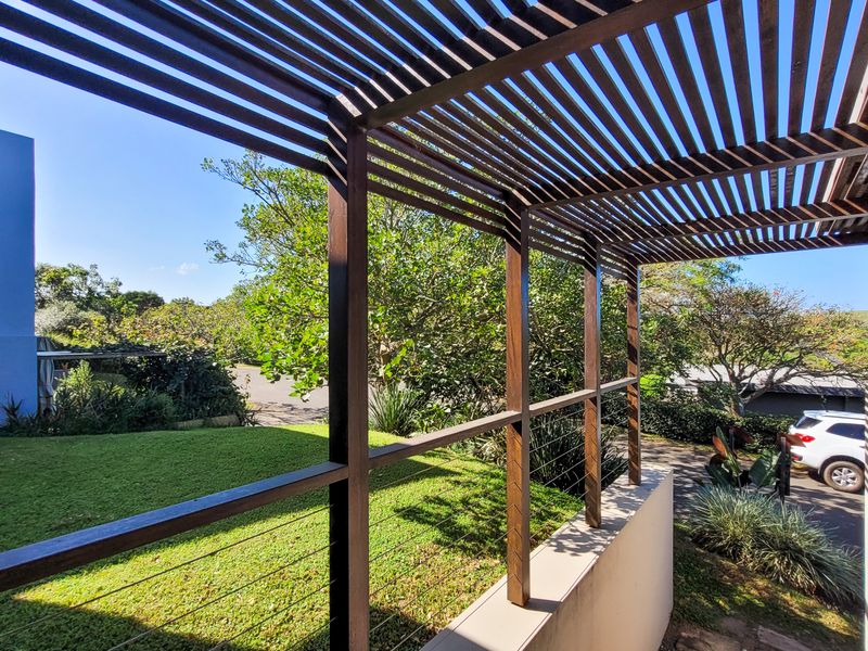 A covered outdoor patio featuring a timber pergola structure and cable railing, overlooking a landscaped garden area.