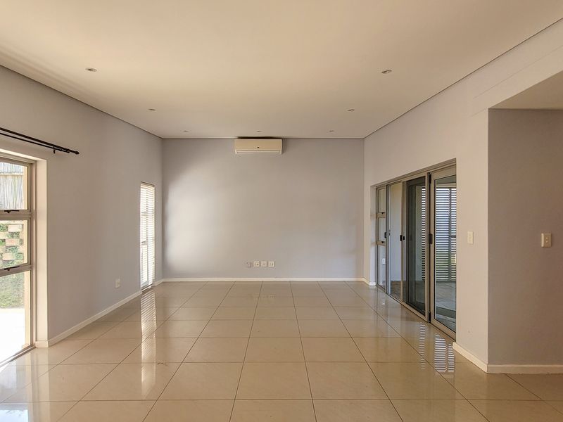 A spacious, light-filled living area featuring neutral tiled flooring, recessed lighting, wall-mounted air conditioning, and large sliding glass doors leading to the exterior.
