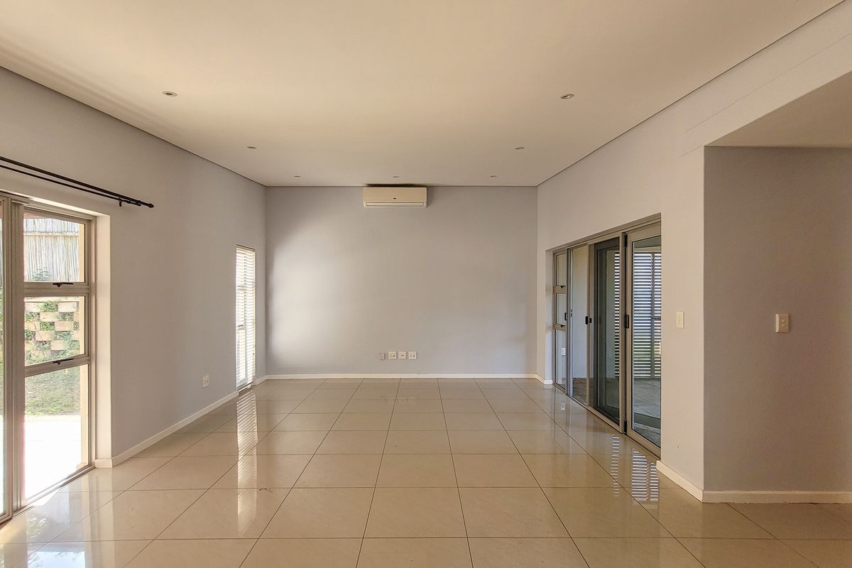 A spacious, light-filled living area featuring neutral tiled flooring, recessed lighting, wall-mounted air conditioning, and large sliding glass doors leading to the exterior.