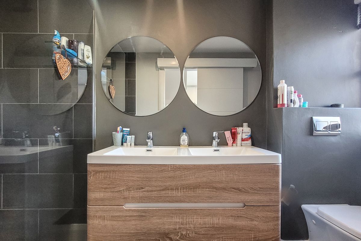 Modern bathroom featuring a double vanity with wood-finish cabinetry, dual circular mirrors, and dark wall tiling.