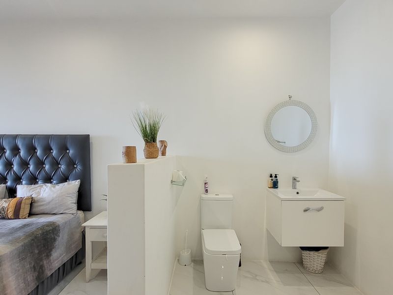 A modern bedroom featuring an open-plan ensuite layout with a wall-mounted vanity, contemporary toilet, and neutral tiled flooring.