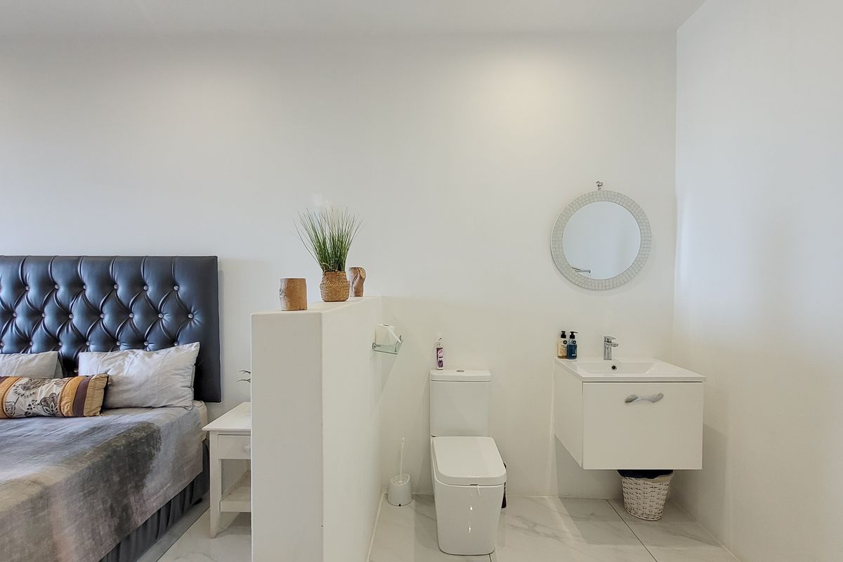 A modern bedroom featuring an open-plan ensuite layout with a wall-mounted vanity, contemporary toilet, and neutral tiled flooring.