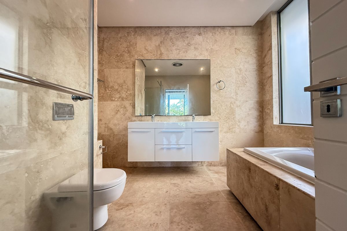 Modern bathroom featuring a double vanity, wall-mounted toilet, glass-enclosed shower, and a built-in bathtub, all finished with elegant travertine tiling.