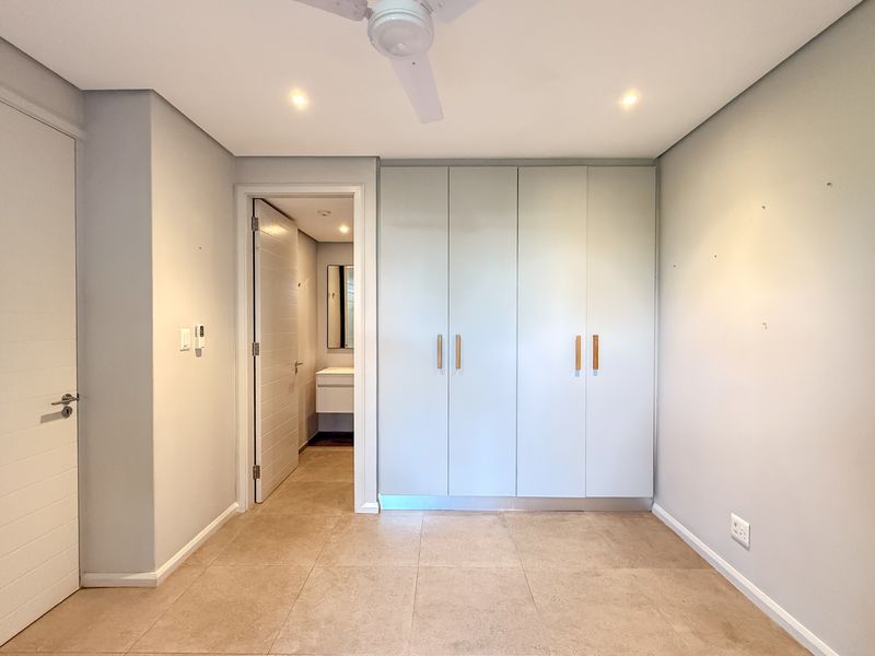A bright, empty bedroom featuring neutral tiled flooring, built-in cupboards, a ceiling fan, and direct access to an en-suite bathroom.