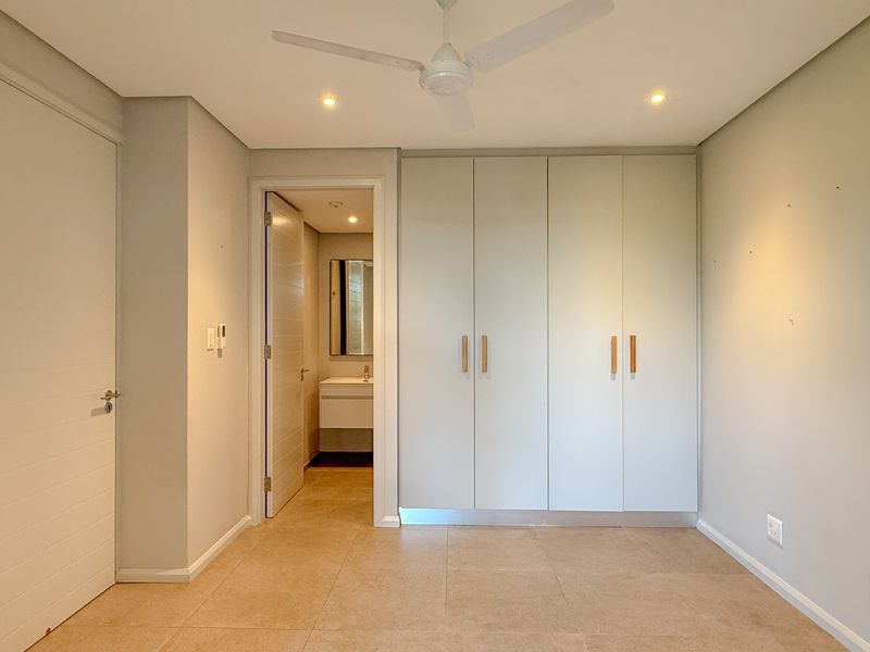 A clean, modern bedroom featuring neutral tiled floors, built-in cupboards, a ceiling fan, and direct access to an en-suite bathroom.