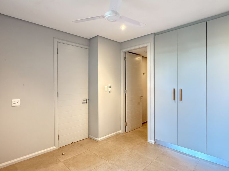 A clean, empty bedroom featuring neutral tiled flooring, built-in cupboards, and a ceiling fan, offering a versatile space ready for personalization.