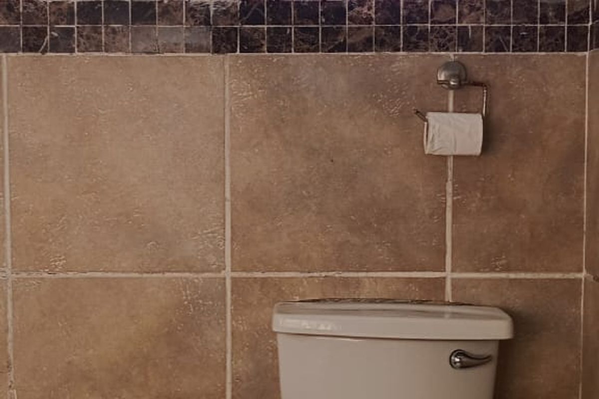 A functional bathroom featuring a standard toilet, pedestal basin, and tiled walls with decorative border accents and security-barred windows.