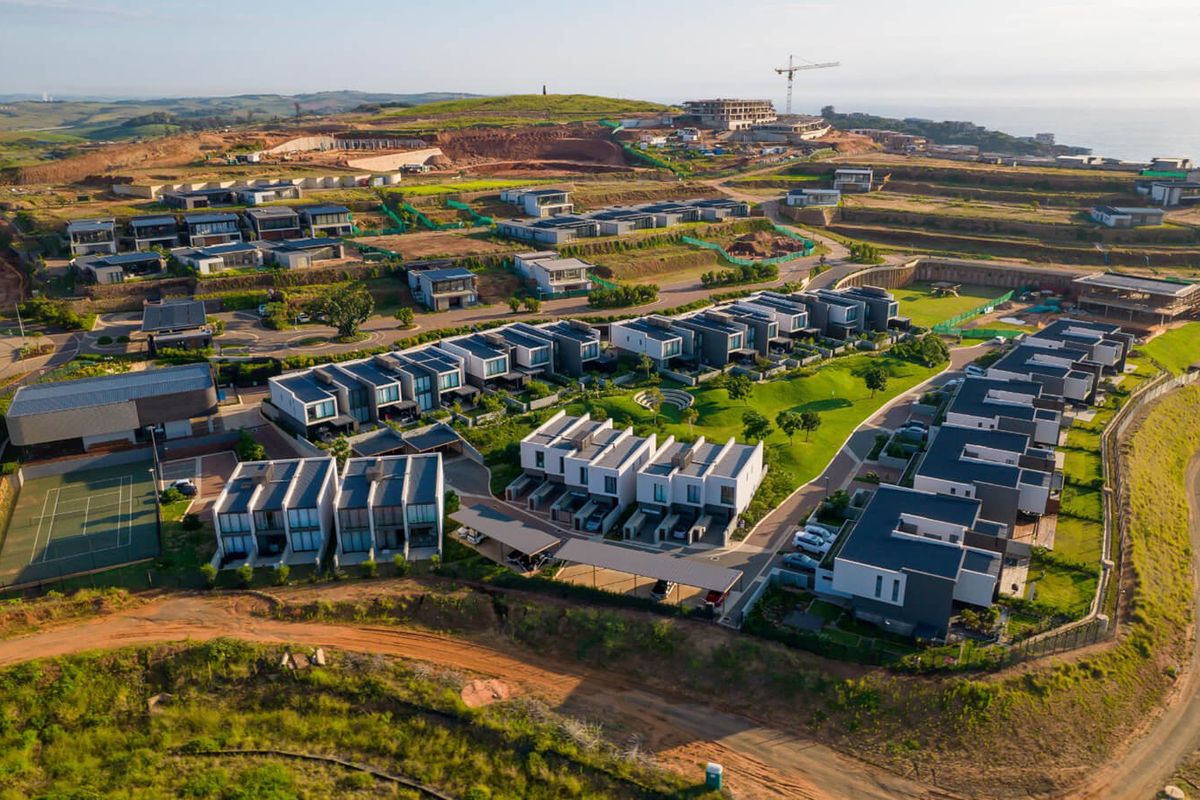 Aerial view of a modern residential estate featuring contemporary townhouses, landscaped green spaces, and a tennis court within a developing coastal community.