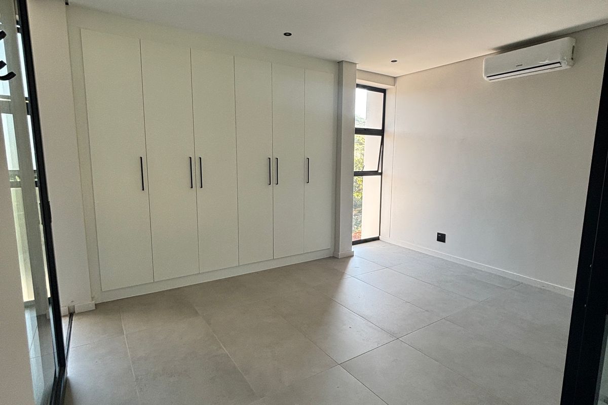 A bright, modern bedroom featuring large format floor tiles, floor-to-ceiling built-in cupboards, and a wall-mounted air conditioning unit.
