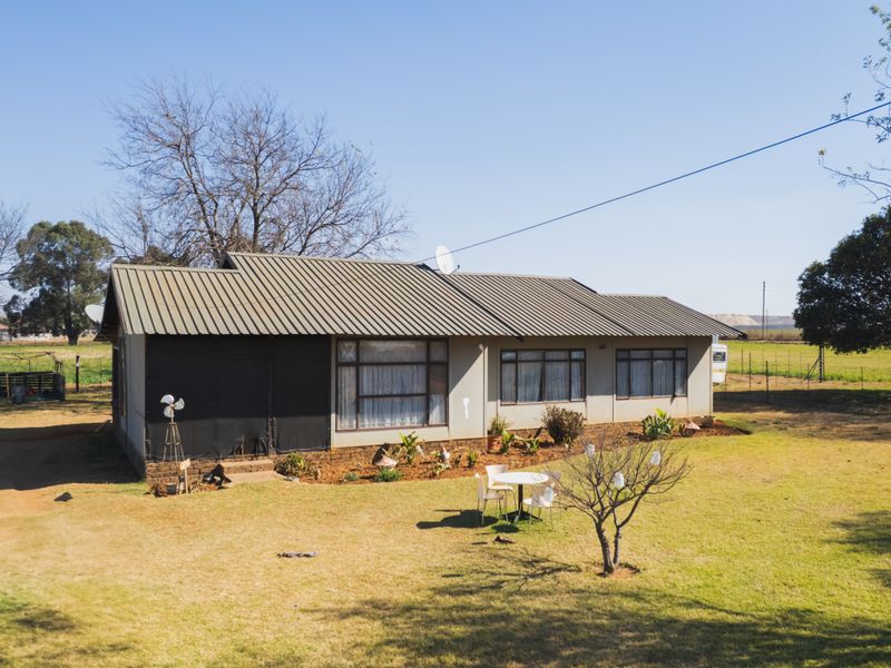 An aerial view of a single-story farm house featuring a metal roof, large windows, and a surrounding garden area on a rural property.