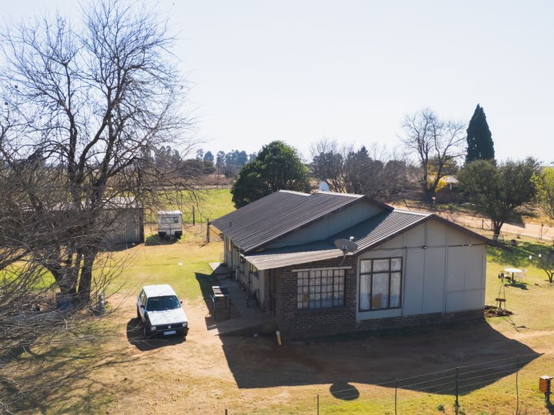 Aerial view of a rural farm property featuring a single-story dwelling with a metal roof, set on a spacious plot with mature trees.