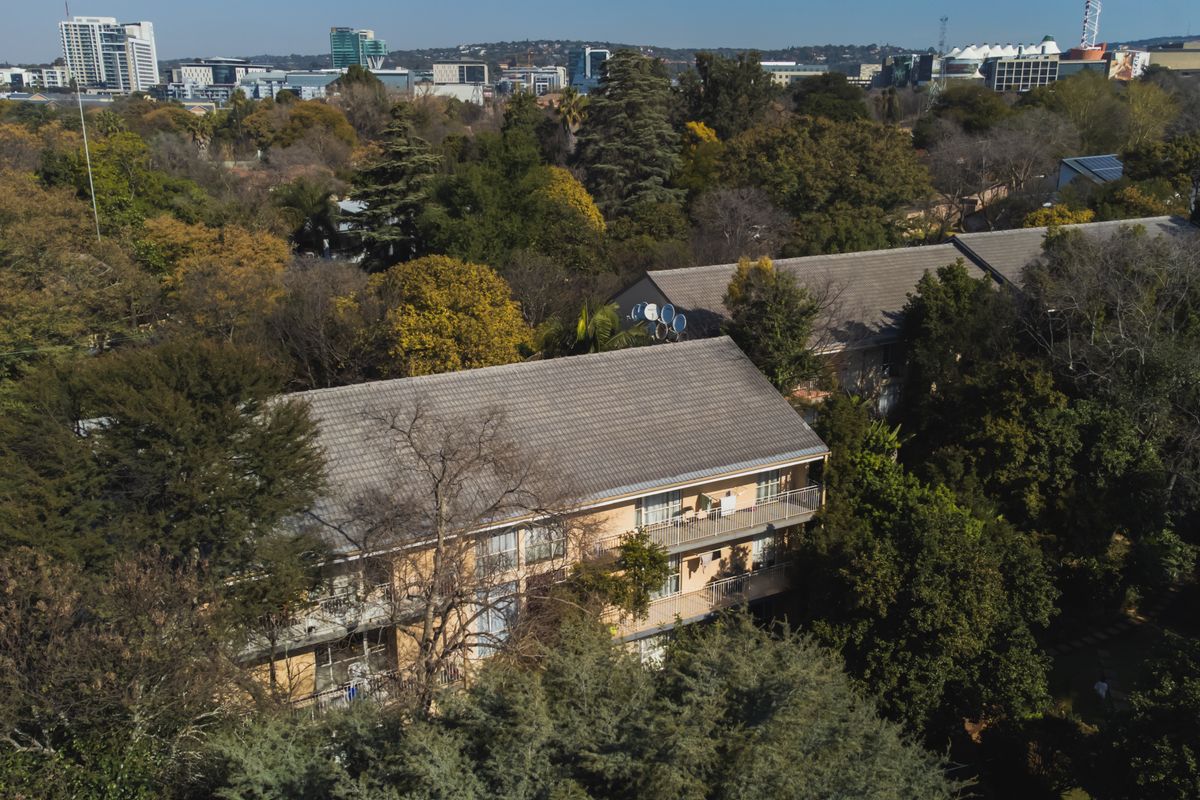 Aerial view of a multi-story apartment complex surrounded by mature trees, featuring tiled roofs and private balconies.