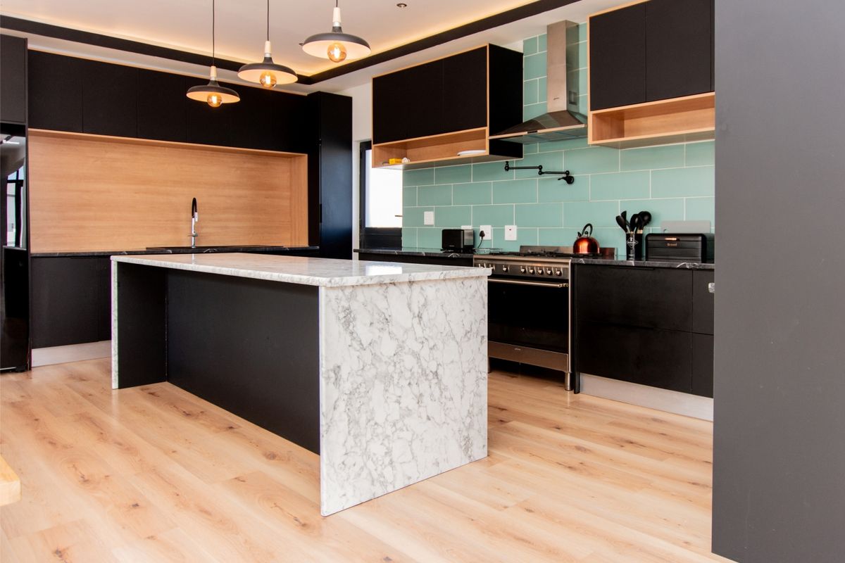 Modern kitchen featuring a marble-topped island, sleek black cabinetry, light wood flooring, and a stylish teal subway tile backsplash.