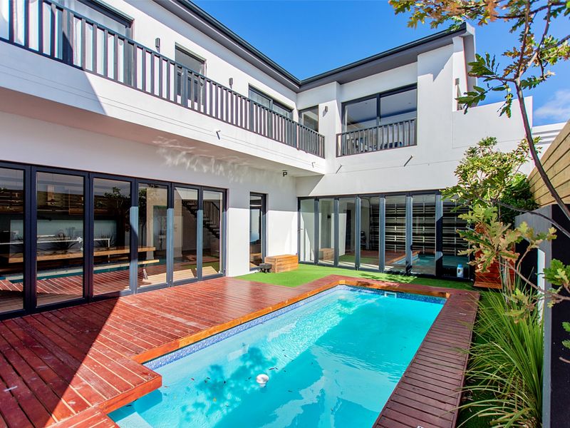 Modern courtyard featuring a swimming pool with timber decking, surrounded by bi-fold doors and a two-story architectural facade with a balcony.