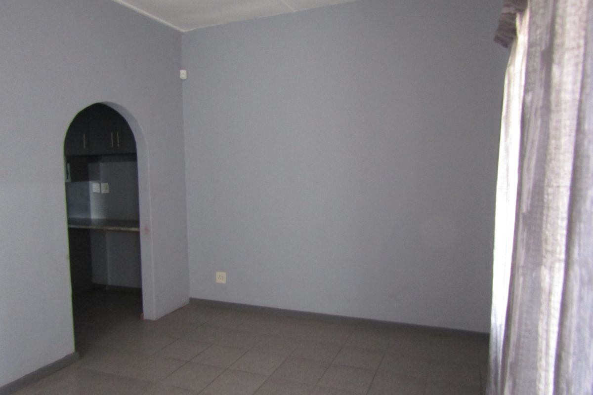 An empty living room featuring neutral grey walls, tiled flooring, and an arched entryway leading to an adjacent kitchen area.