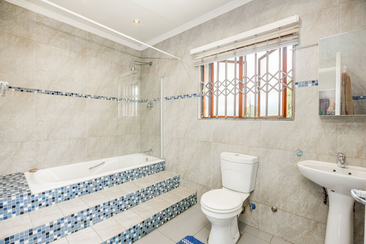 A well-maintained bathroom featuring a raised built-in bathtub with a glass screen, pedestal basin, toilet, and neutral wall tiling with decorative mosaic accents.