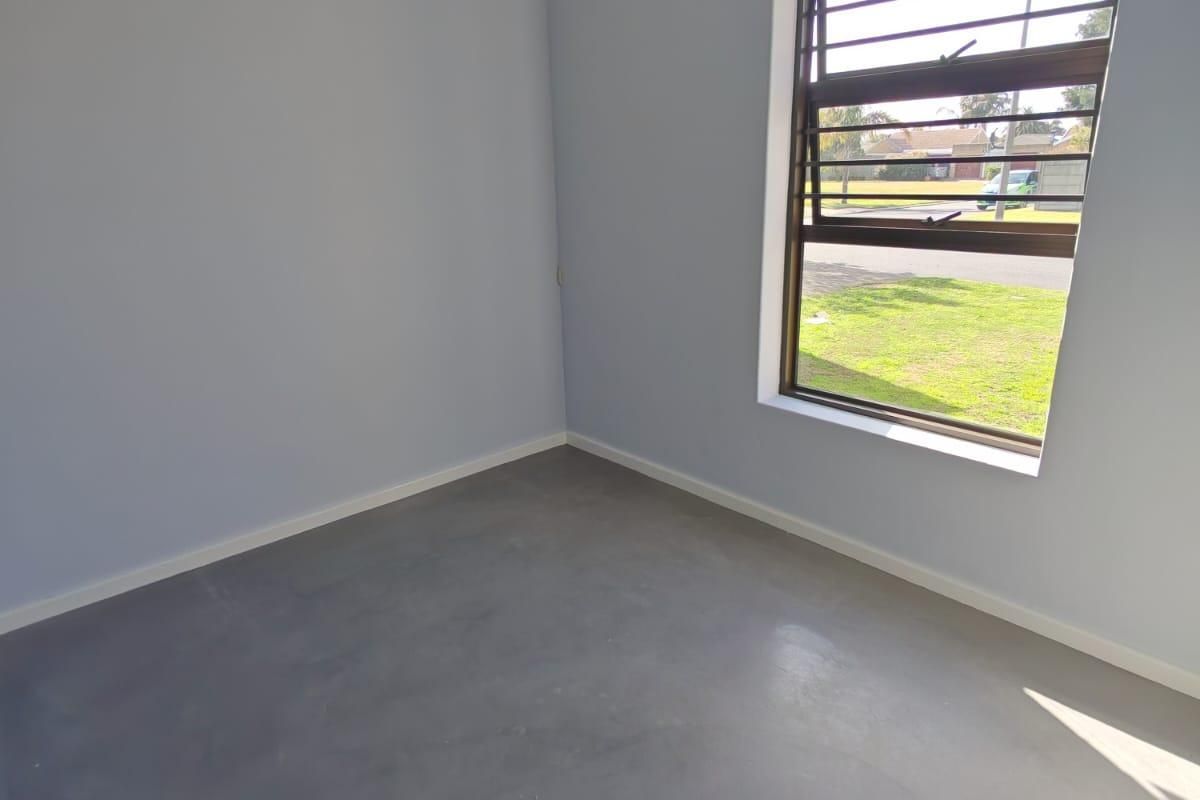 A bright, empty bedroom featuring polished concrete flooring, coving, and a window fitted with security bars.