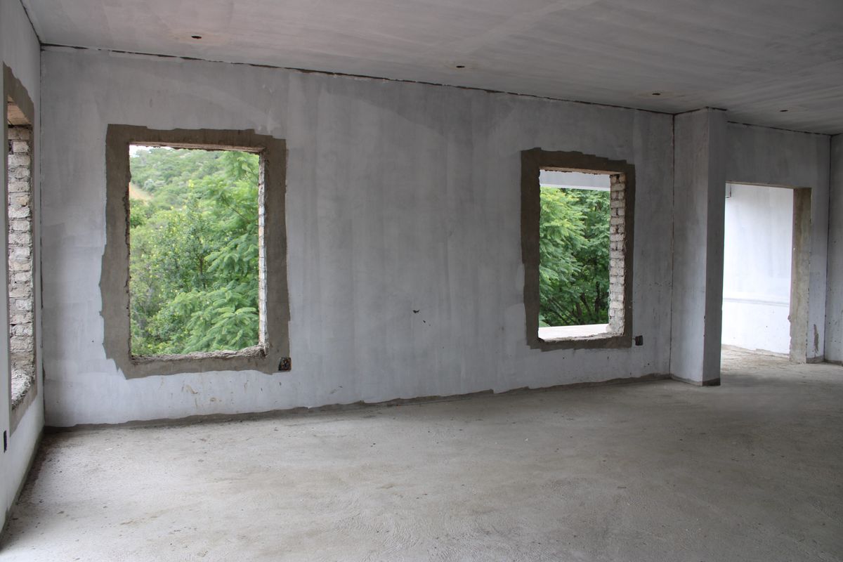 An unfinished interior space in shell condition, featuring exposed concrete flooring, plastered walls, and open window apertures overlooking greenery.