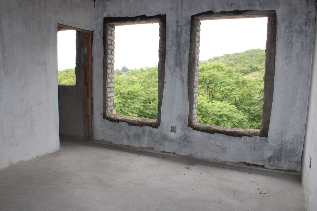 An unfinished interior room featuring exposed concrete walls and flooring with large open window apertures overlooking a lush green landscape.
