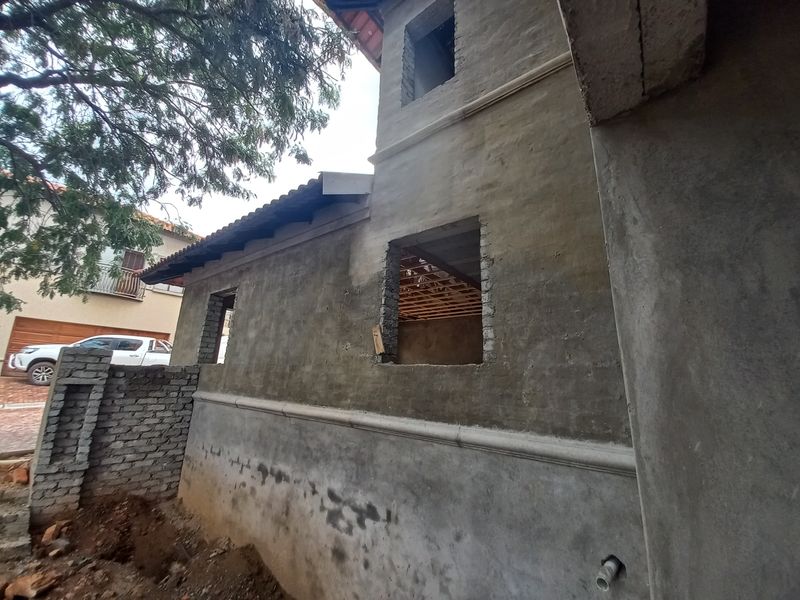 Exterior view of a house under construction, featuring unfinished plaster walls, exposed brickwork, and missing window frames, indicating a project in progress.