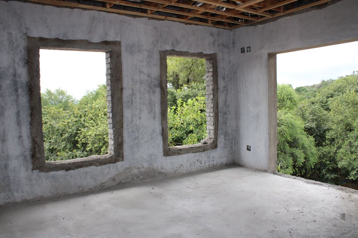An unfinished interior room in shell condition, featuring exposed ceiling joists, raw concrete walls, and open window and door apertures overlooking greenery.