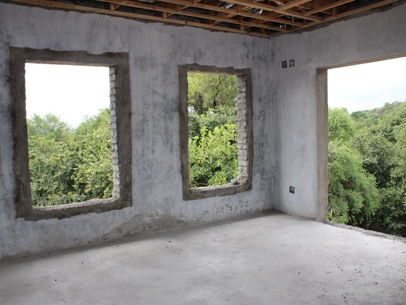 An unfinished interior room in shell condition, featuring exposed ceiling joists, raw concrete walls, and open window and door apertures overlooking greenery.