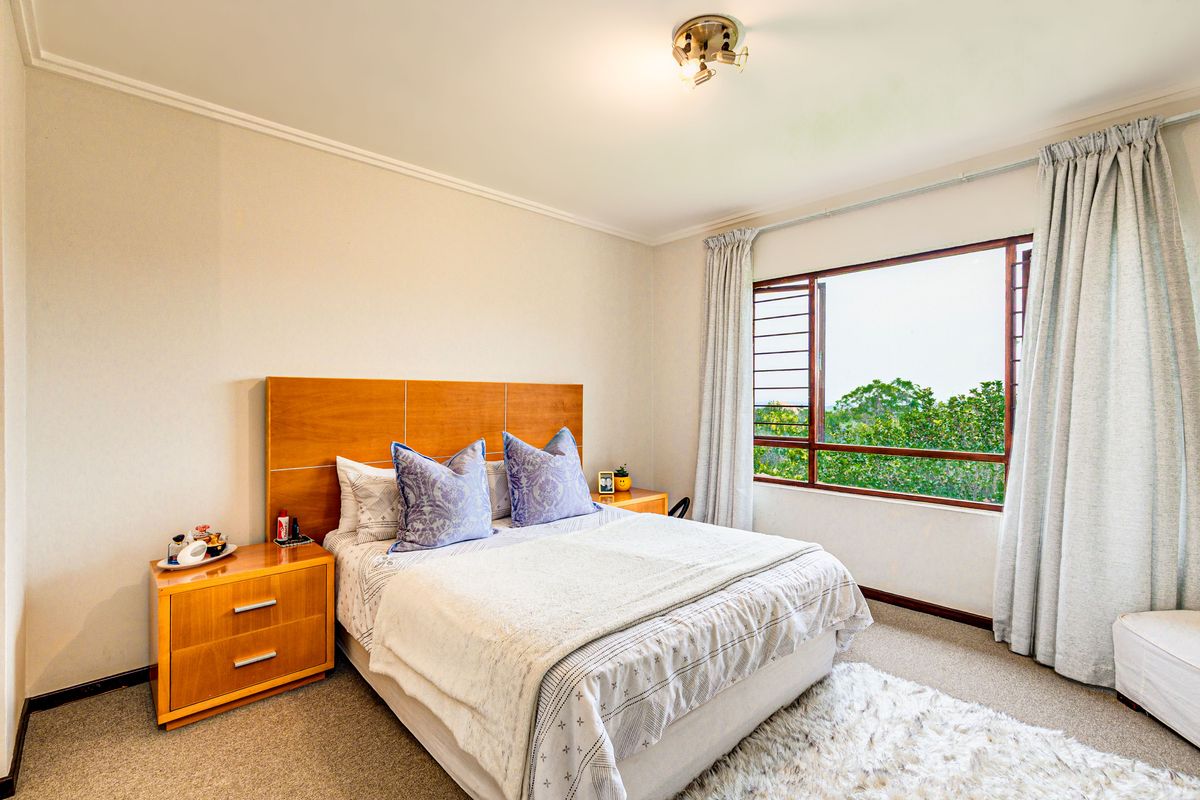 A well-lit bedroom featuring a large window with security bars, neutral carpeting, and a ceiling-mounted light fixture.