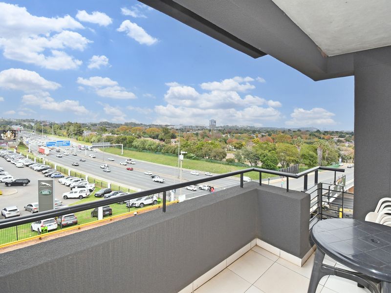 A private balcony featuring tiled flooring, a modern dark grey finish, and a metal railing, offering expansive views over the surrounding area and highway.