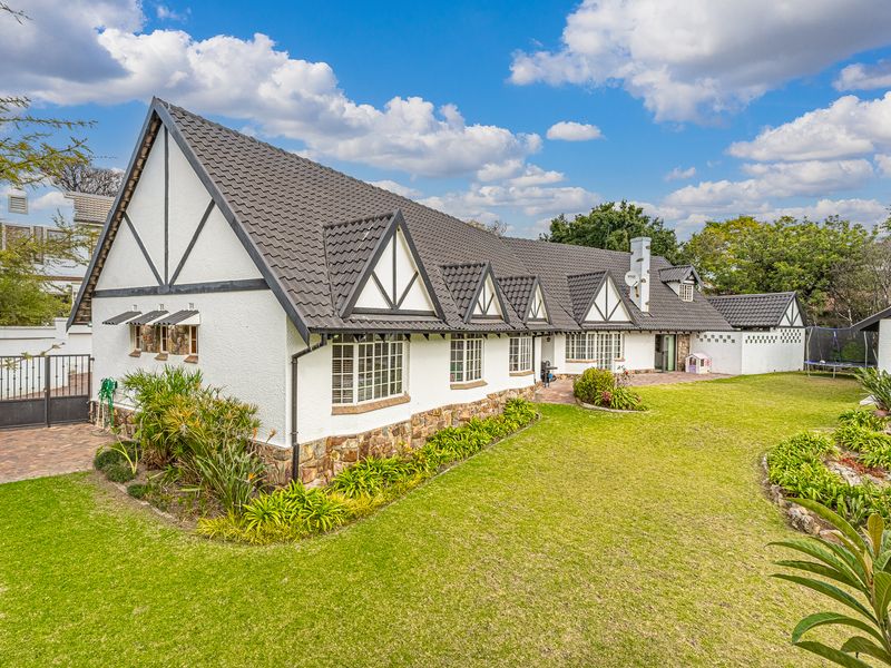 A well-maintained Tudor-style house featuring a dark tiled roof, stone base detailing, and a spacious landscaped garden with a paved patio area.