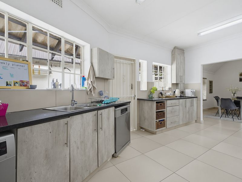 A functional kitchen featuring light wood-look cabinetry, dark countertops, stainless steel sink, and tiled flooring, with an adjacent dining area visible.