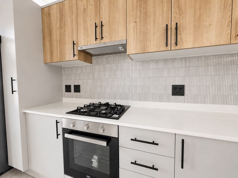 Modern kitchen featuring light wood-tone upper cabinets, white lower cabinetry, stone countertops, a gas hob, and a sleek tiled backsplash.