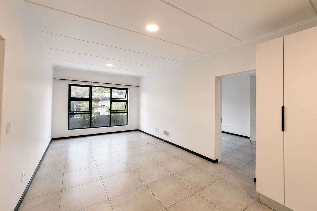 A bright, empty bedroom featuring neutral floor tiles, recessed ceiling lighting, and a built-in wardrobe, with a view through the window.
