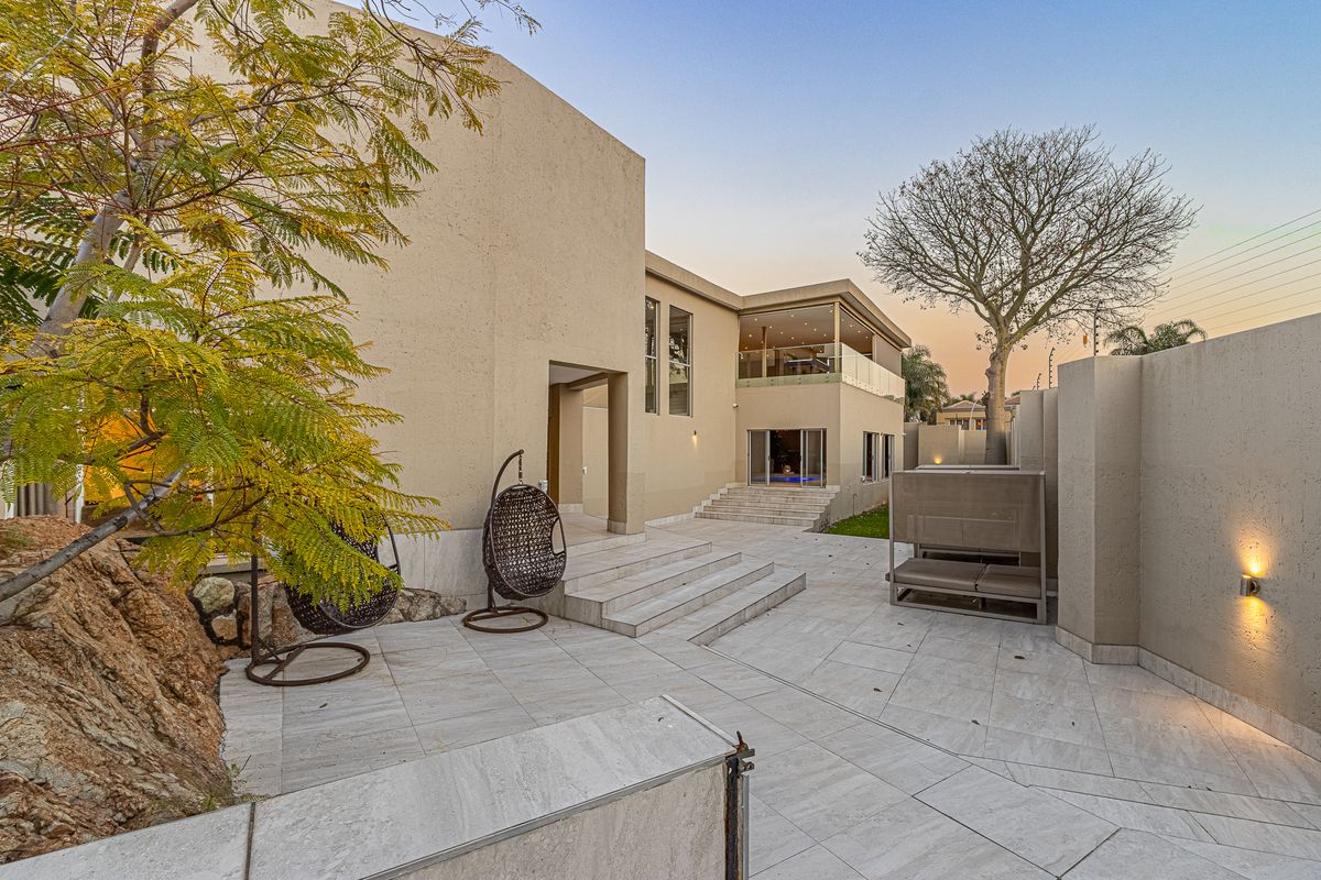 A modern rear exterior featuring a spacious tiled patio, stone retaining wall, and a balcony with glass balustrades overlooking the garden area.