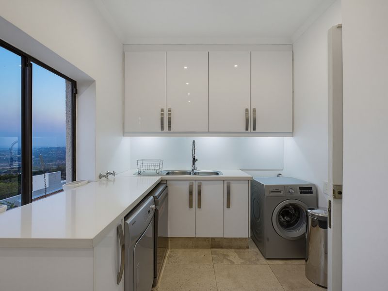A modern, well-lit laundry room featuring sleek white cabinetry, stone countertops, an integrated sink, and tiled flooring with a scenic window view.