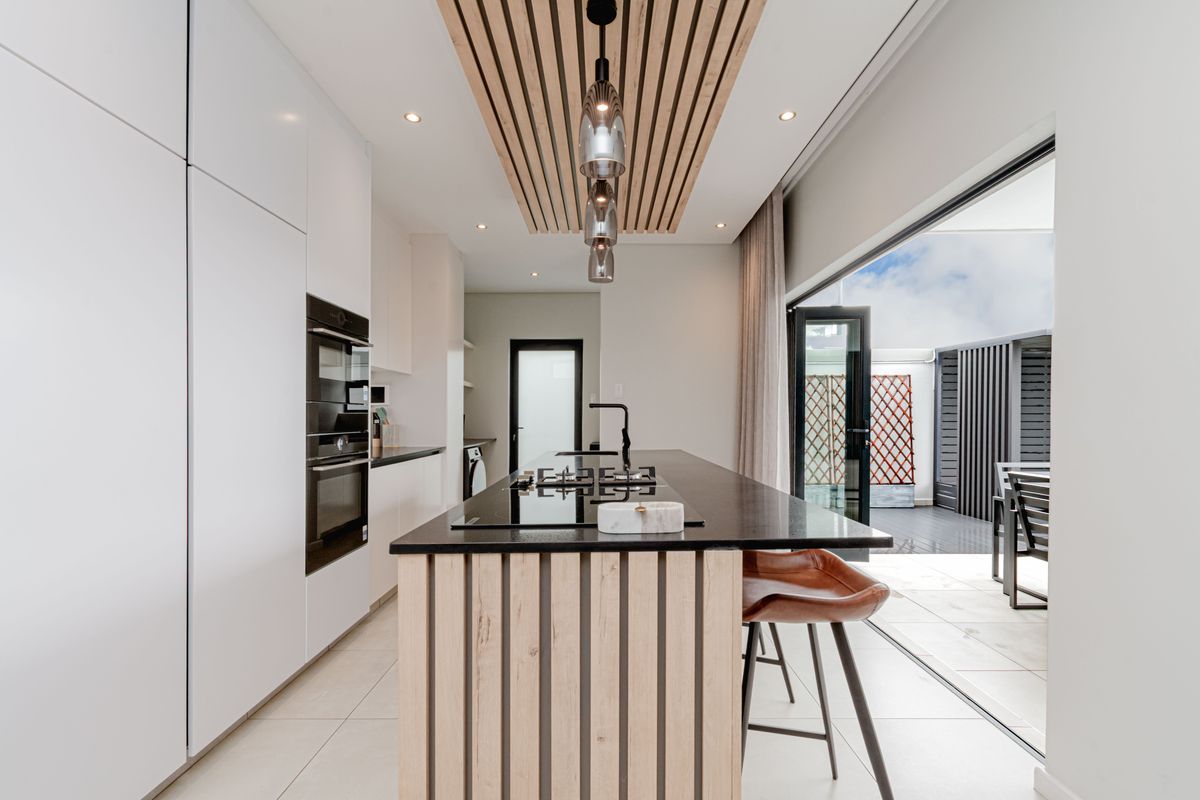 Modern kitchen featuring a central island with a gas hob, built-in appliances, and a stylish slatted timber ceiling detail, opening onto an outdoor patio area.