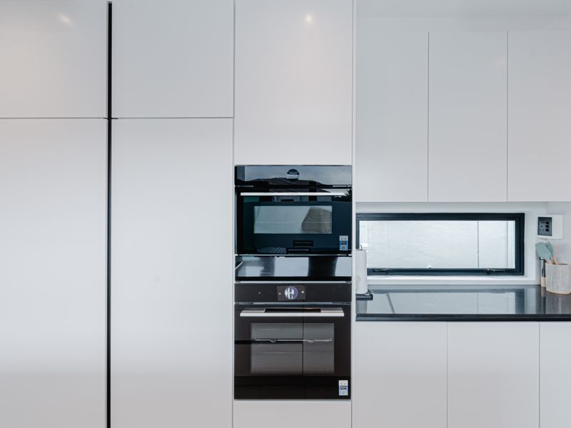 Modern kitchen featuring sleek white high-gloss cabinetry, integrated black oven and microwave, and a contrasting black granite countertop with a horizontal window.