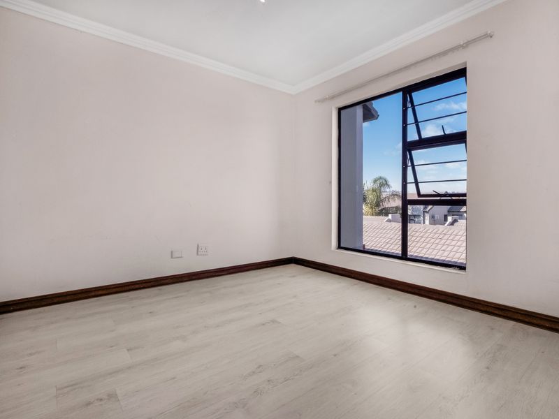 A bright, empty bedroom featuring light-toned laminate flooring, a built-in wardrobe, and a large window with views of the surrounding neighborhood.