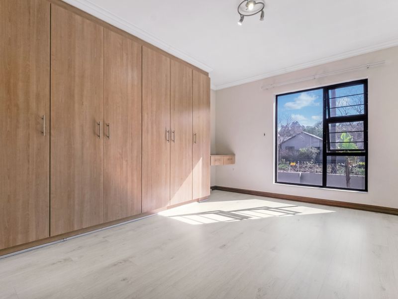 A bright, empty bedroom featuring light-toned laminate flooring, extensive built-in wooden cupboards, and a large window with an aluminium frame.
