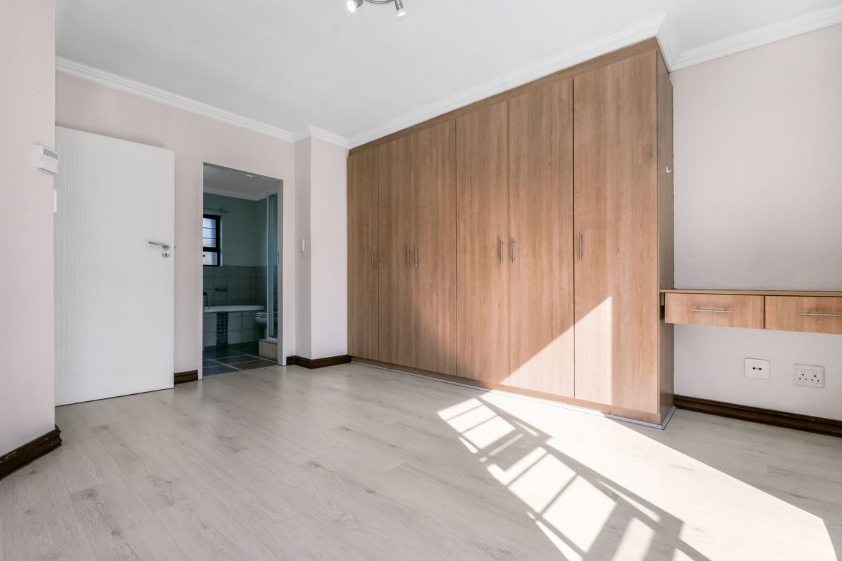 A bright, empty bedroom featuring light laminate flooring, extensive built-in wooden cupboards, and an integrated desk, with an adjoining bathroom visible.