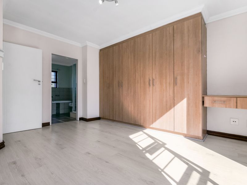 A bright, empty bedroom featuring light laminate flooring, extensive built-in wooden cupboards, and an integrated desk, with an adjoining bathroom visible.