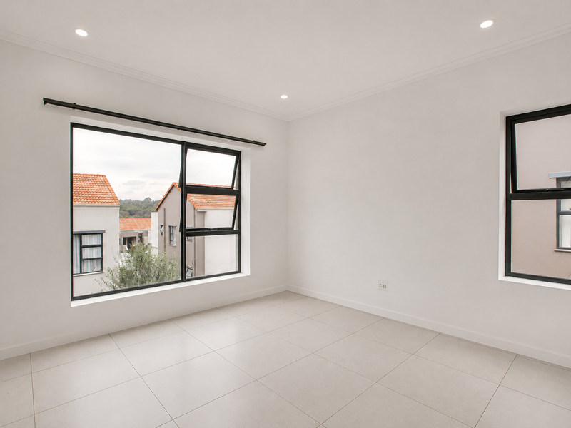 A bright, empty bedroom featuring neutral tiled flooring, recessed downlights, and large black aluminium-framed windows offering a view of the surrounding complex.